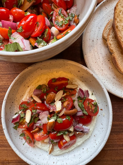  A plate of whipped ricotta topped with a fresh tomato salad made with cherry tomatoes, red onions, basil, and almonds, served alongside toasted bread slices.